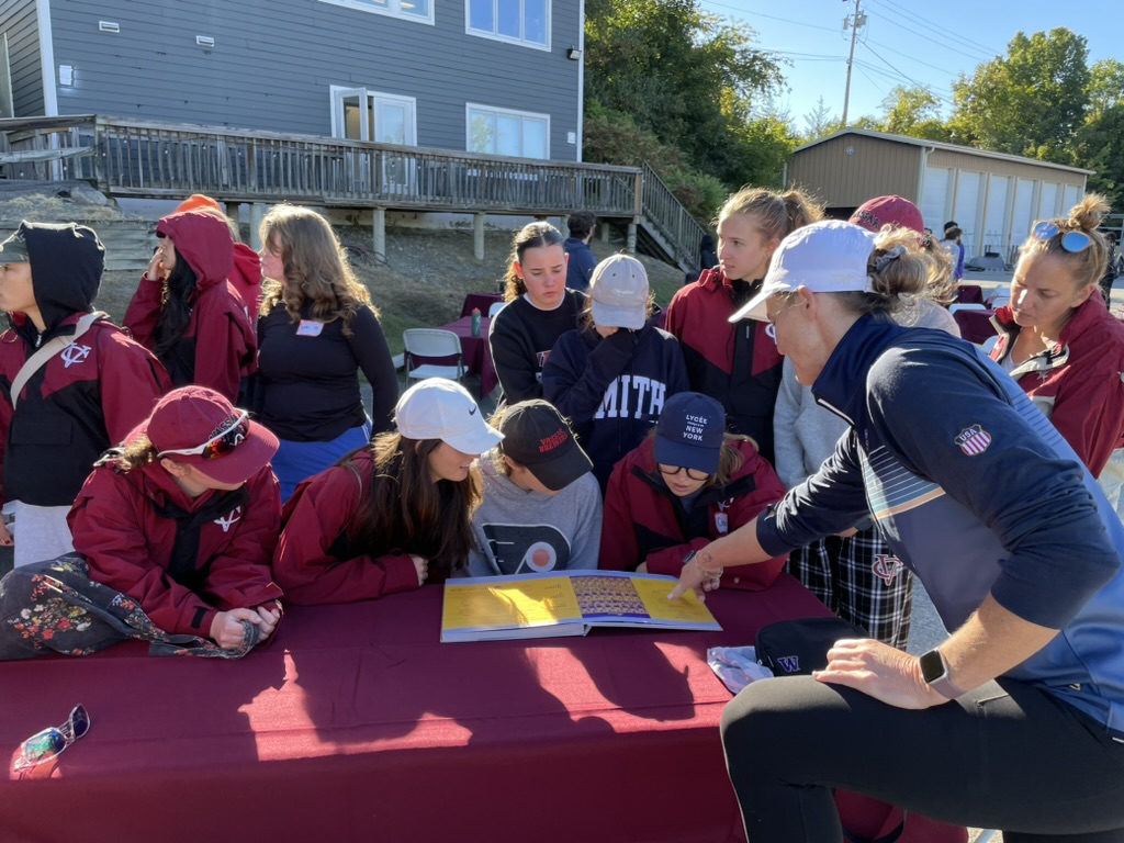 Current team members and an alumna gathered around a table looking at an old photo book outside the boathouse